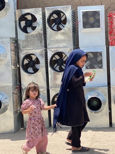 Young girls walk past industrial coolers in Kandahar