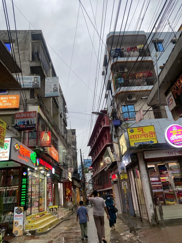 Busy Dhaka street: chaotic wiring overhead, snack stall below