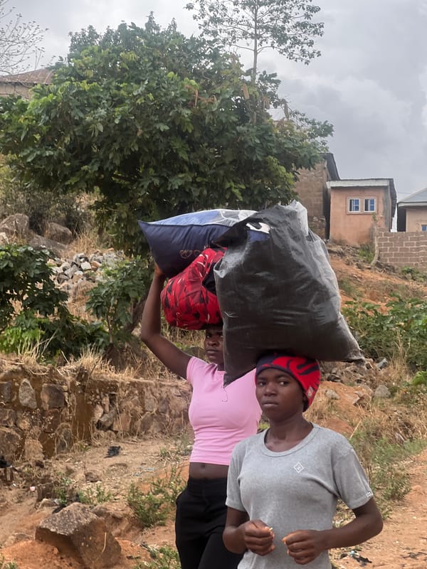 Two people carry large bags on dirt terrain in Jos
