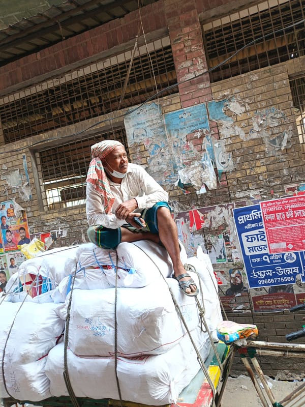 Man in traditional dress sits atop bag pile at Dhaka rickshaw park