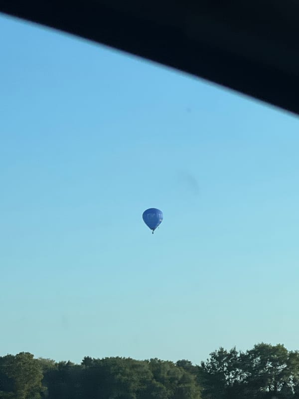 Hot air balloons drift over treetops as a logging truck waits at a red light