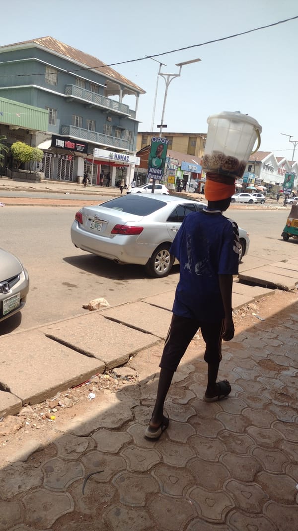 Person carries container on head along Jos sidewalk