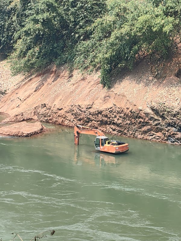 Excavator submerged in river as man sits on road