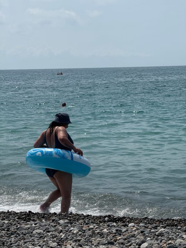 Busy resort beach day with swimmers, sunbathers, and a hilltop cross