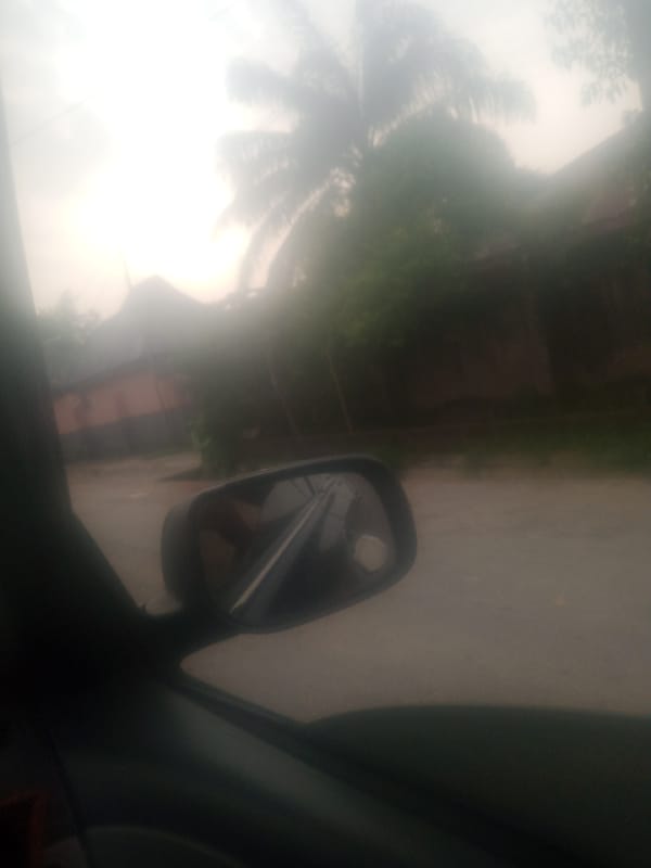 Street-level drive through hazy Jalingo with auto-rickshaw in view
