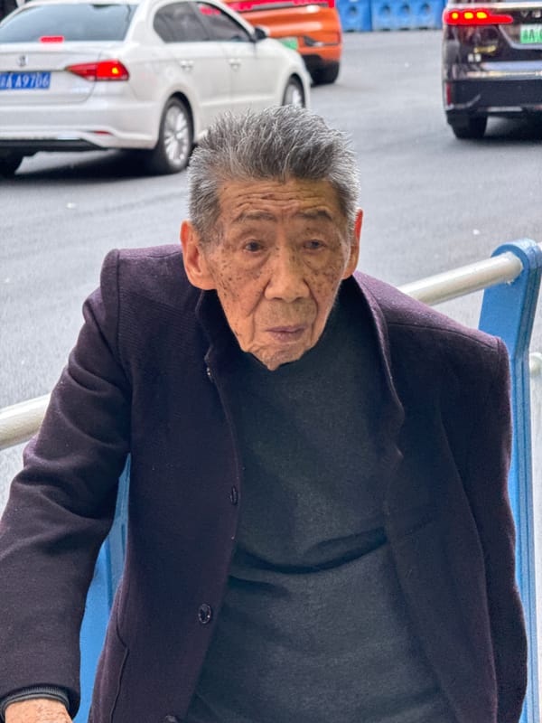 Elderly man sits, people gather near poster-covered building