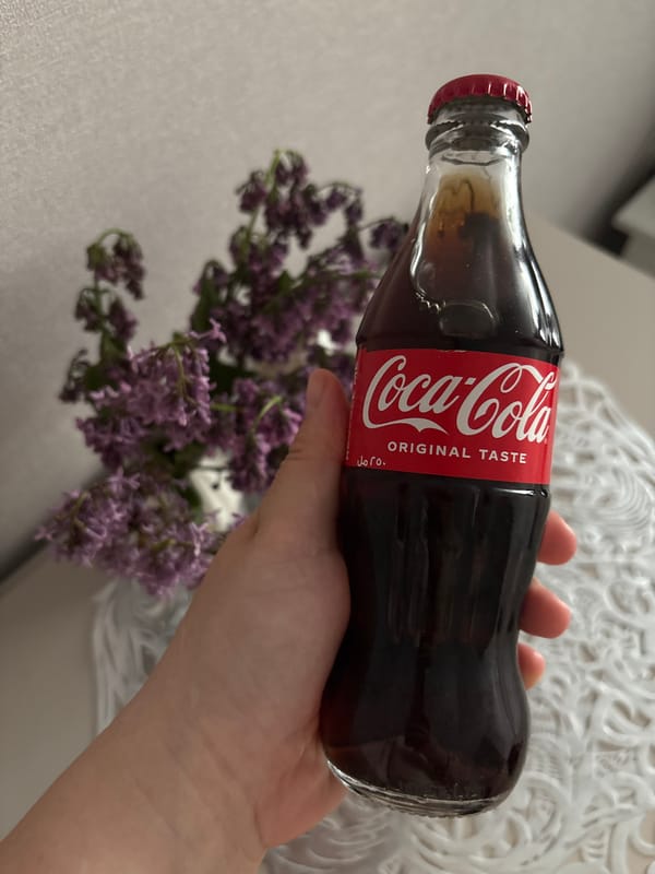 Person holds tilted Coca-Cola bottle above purple flowers