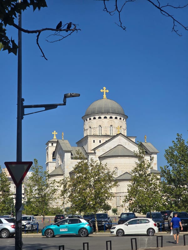 Street scene outside a white Orthodox church with parked cars and flowering trees