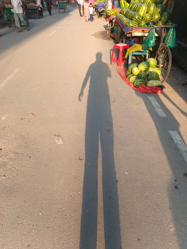 Man carries bag past watermelon stall in sunny Dhaka