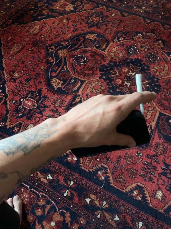 Tattooed hand holds cigarette and phone over patterned rug