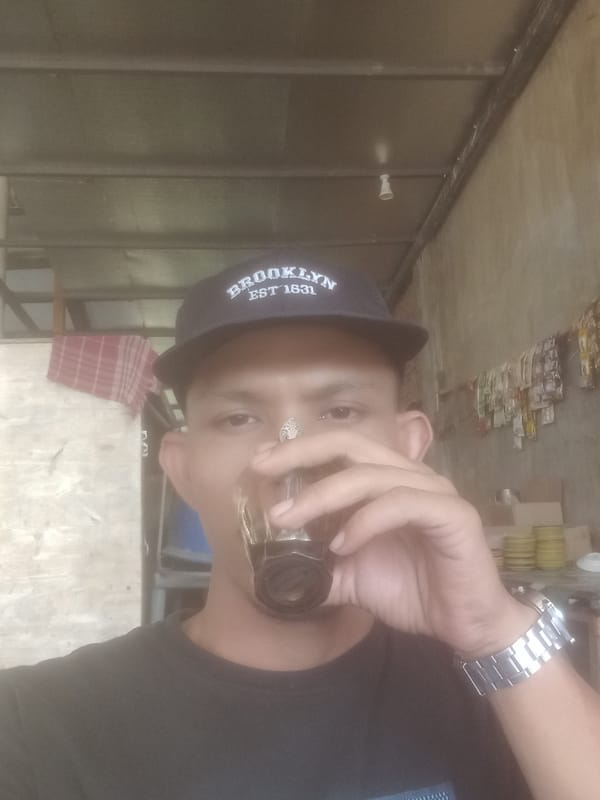 Sipping a dark drink in Keudejungkagajah, Brooklyn cap on