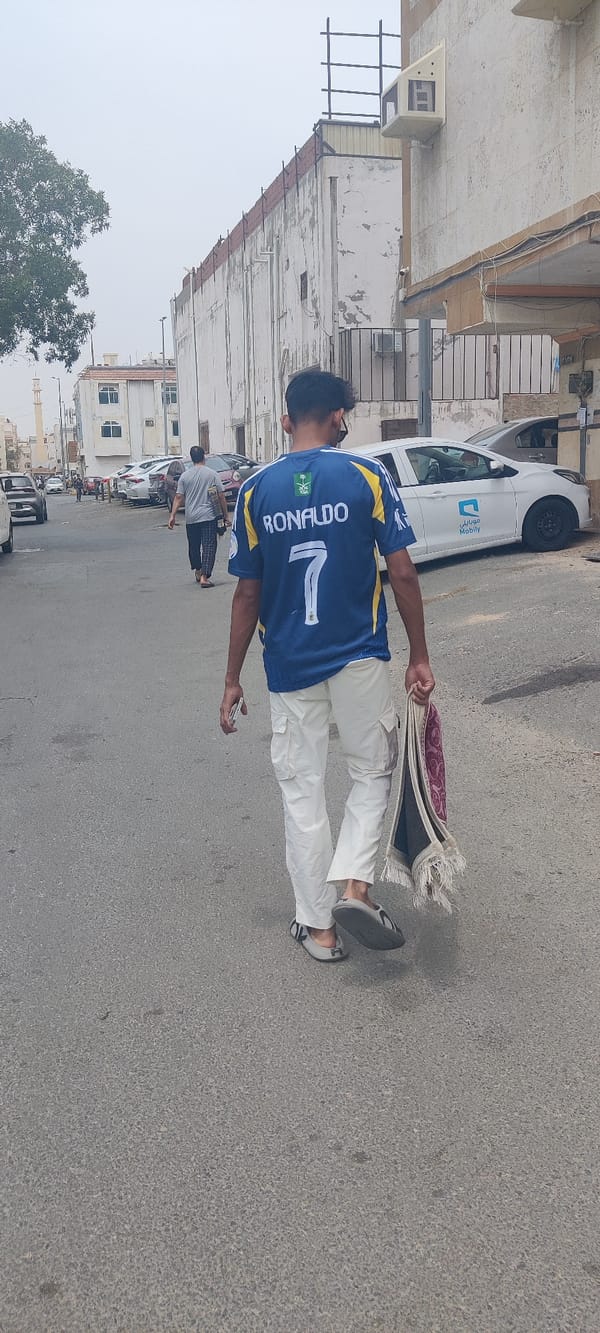 Pedestrians walk Jeddah street, man carries prayer rug