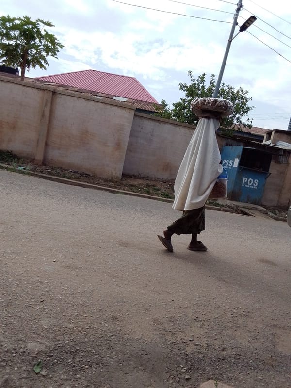 Person in white cloth carries head tray on urban street