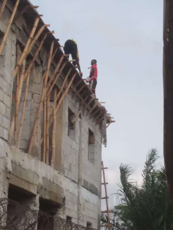 Concrete block building mid-construction, workers active on scaffolding and roof