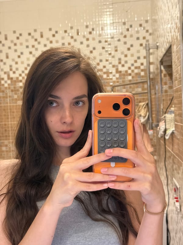 Bathroom mirror selfie with orange phone, grey tank top