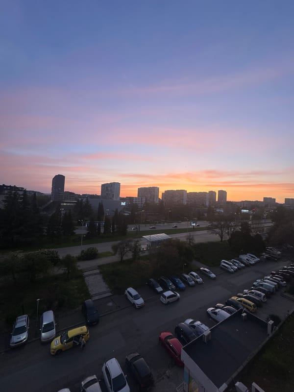 Urban dusk scene shows apartment buildings, parked cars, greenery