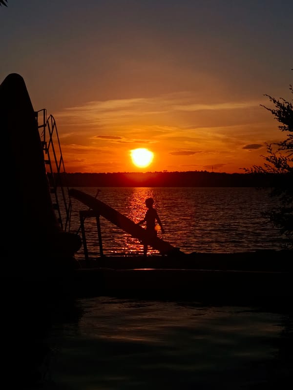 Lakeside piano set at sunset, robed performer on wooden stage.